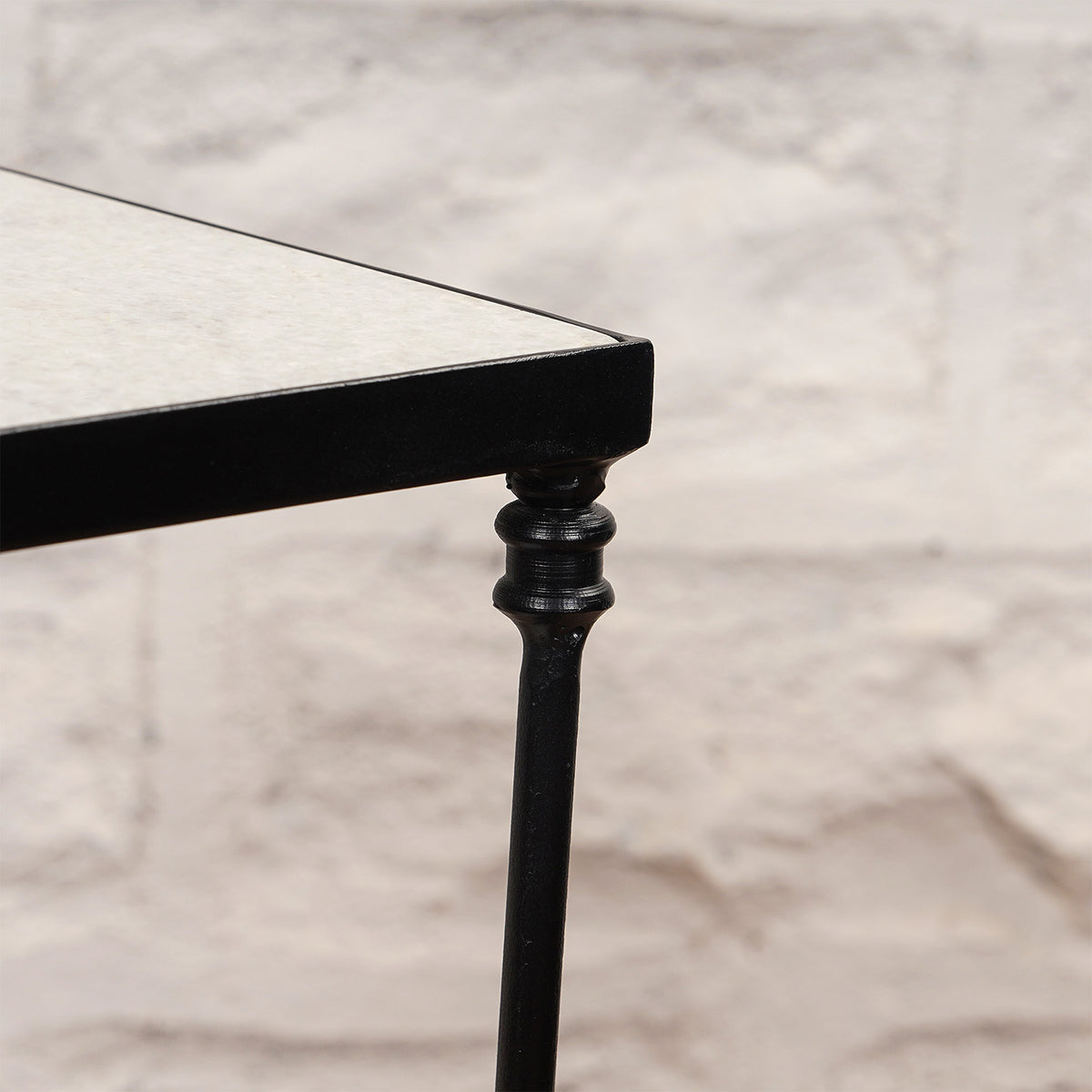 Close-up of a black metal leg with a decorative design on the John Robshaw Veda Side Table, supporting its rectangular tabletop against a light stone wall background, showcasing the minimalist elegance characteristic of this iron side table.