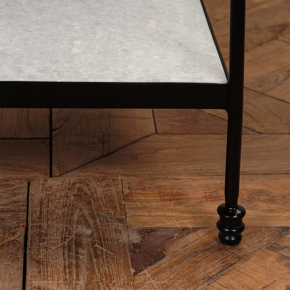 Close-up of the Veda Side Table by John Robshaw, featuring a black iron leg and marble shelf resting on a wooden parquet floor, bringing a minimalist touch to the space.