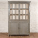 The Kali Teak Cabinet by John Robshaw features glass-paneled upper doors and solid lower doors, showcasing a vintage-inspired design. It is displayed against a white brick wall atop a wooden floor.