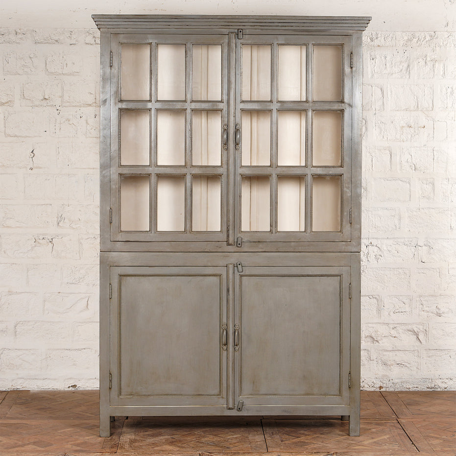 The Kali Teak Cabinet by John Robshaw features glass-paneled upper doors and solid lower doors, showcasing a vintage-inspired design. It is displayed against a white brick wall atop a wooden floor.