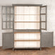 The Kali Teak Cabinet by John Robshaw, featuring a two-section gray vintage design with glass-paneled upper doors and solid lower doors that open to reveal empty white shelves, stands against a white brick wall on a wooden floor.