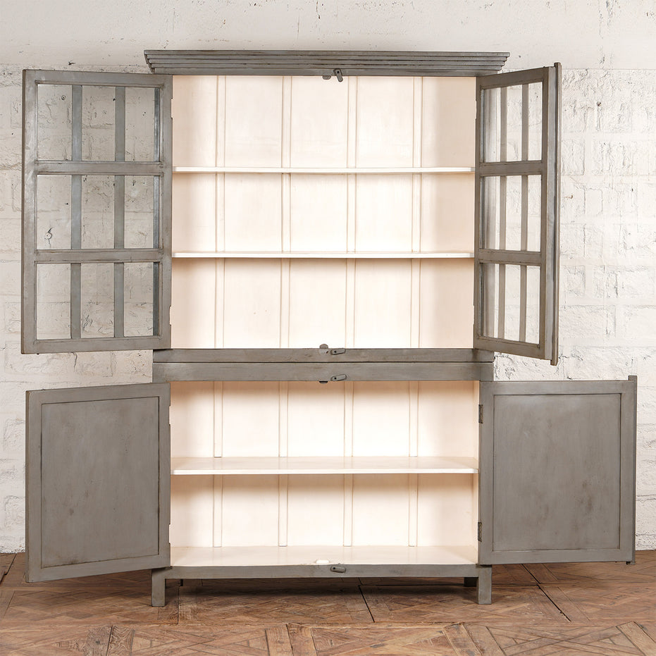 The Kali Teak Cabinet by John Robshaw, featuring a two-section gray vintage design with glass-paneled upper doors and solid lower doors that open to reveal empty white shelves, stands against a white brick wall on a wooden floor.