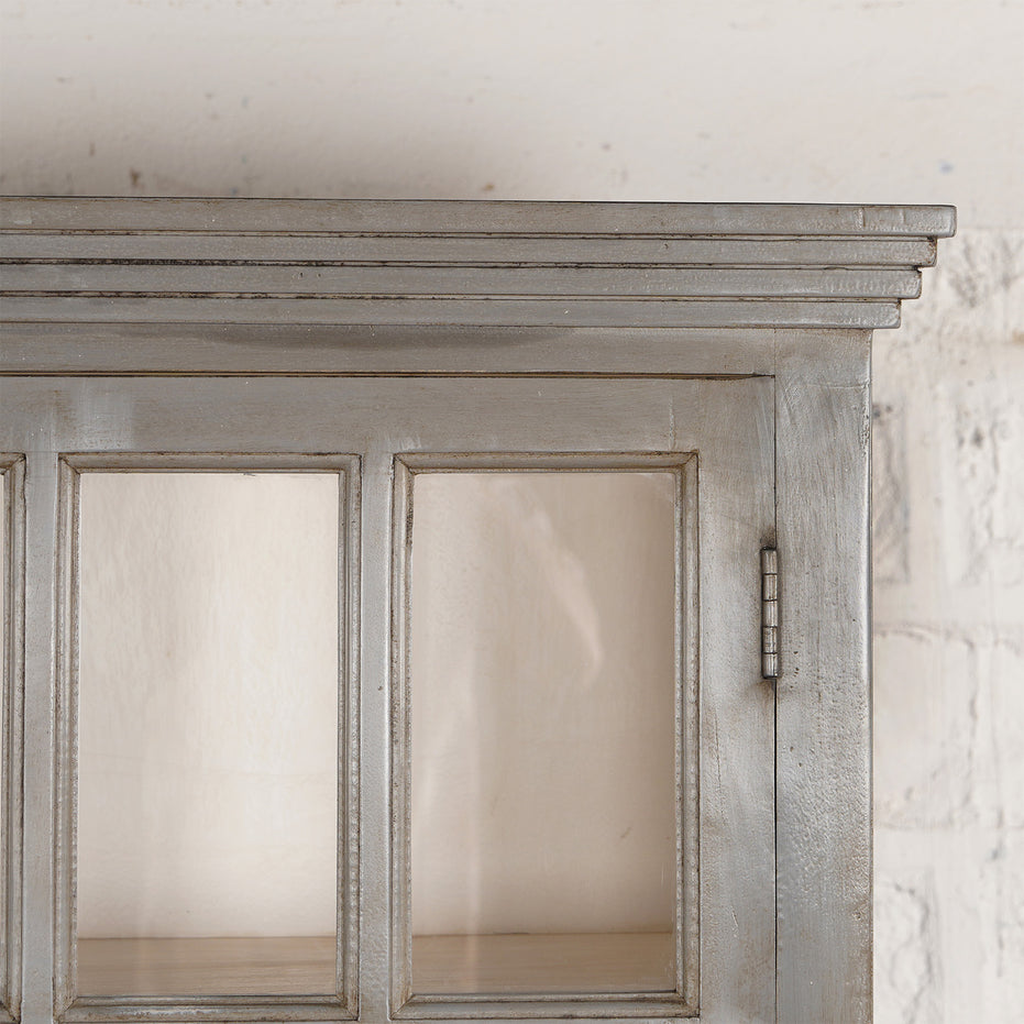 Close-up of the upper portion of the John Robshaw Kali Teak Cabinet, featuring glass doors, decorative trim, and set against a white textured wall background—an elegant nod to vintage furniture style.