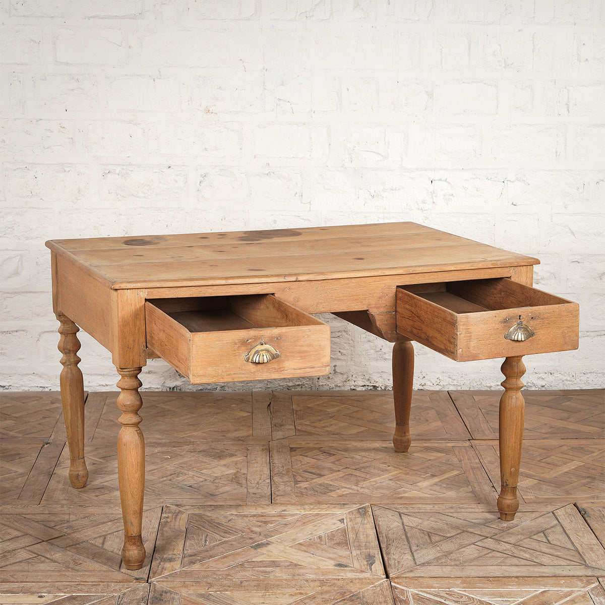 The Smriti Teak Desk by John Robshaw features two open drawers, graceful spindle legs, and a light natural finish. Placed on a patterned wooden floor against a white brick wall, this desk exudes timeless charm and brings elegant style to any space.