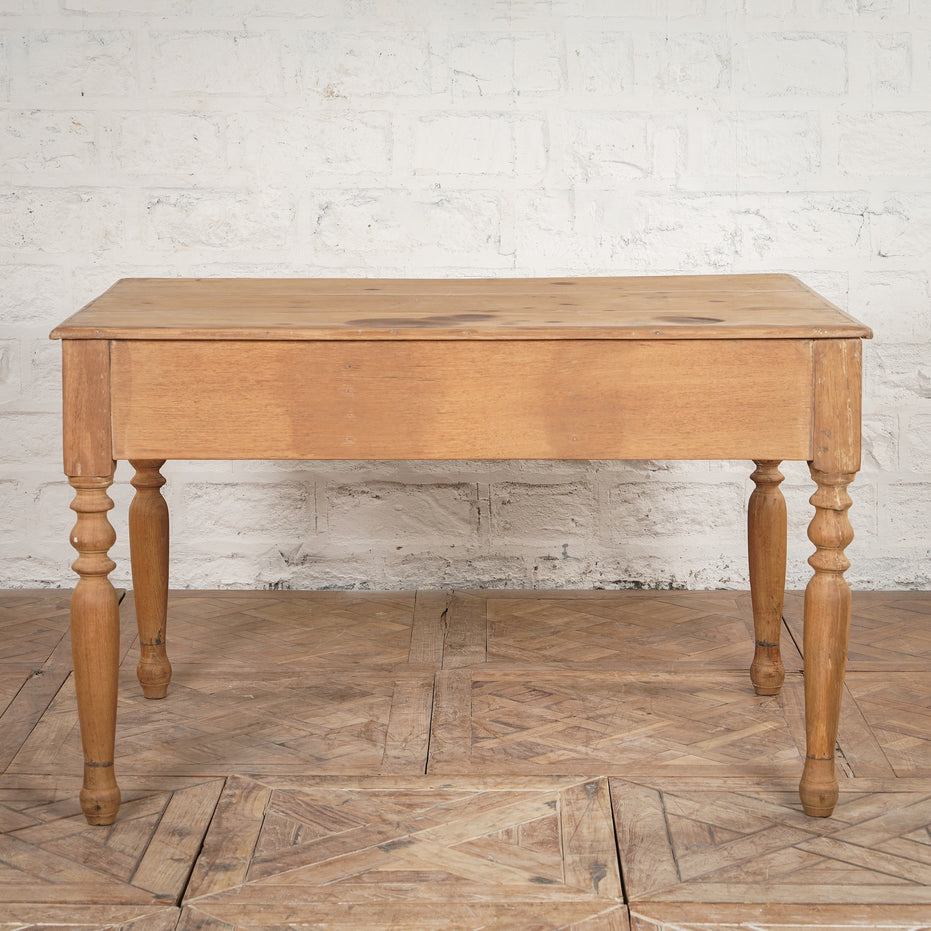 The Smriti Teak Desk by John Robshaw, featuring a rectangular top and four turned legs, stands on a patterned wooden floor against a white brick wall.