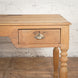 Close-up of the Smriti Teak Desk by John Robshaw, a vintage-inspired writing desk with a single drawer, a distinctive metal shell-shaped handle, turned leg detailing, and charming signs of wear, set against a white brick wall.