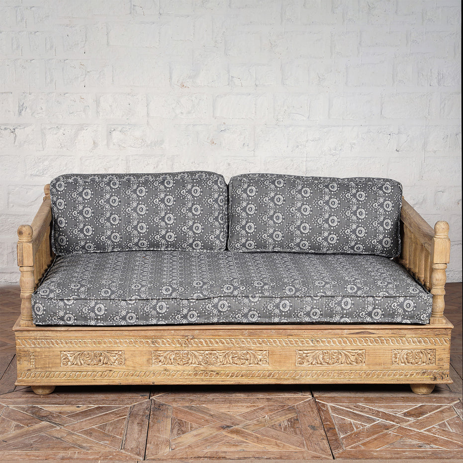 Experience vintage furniture at its finest with the Yashti Wooden Daybed by John Robshaw, featuring a carved wooden base and armrests, complemented by black and white block printed fabric seat and back cushions—beautifully set against a white brick wall on a wooden floor.