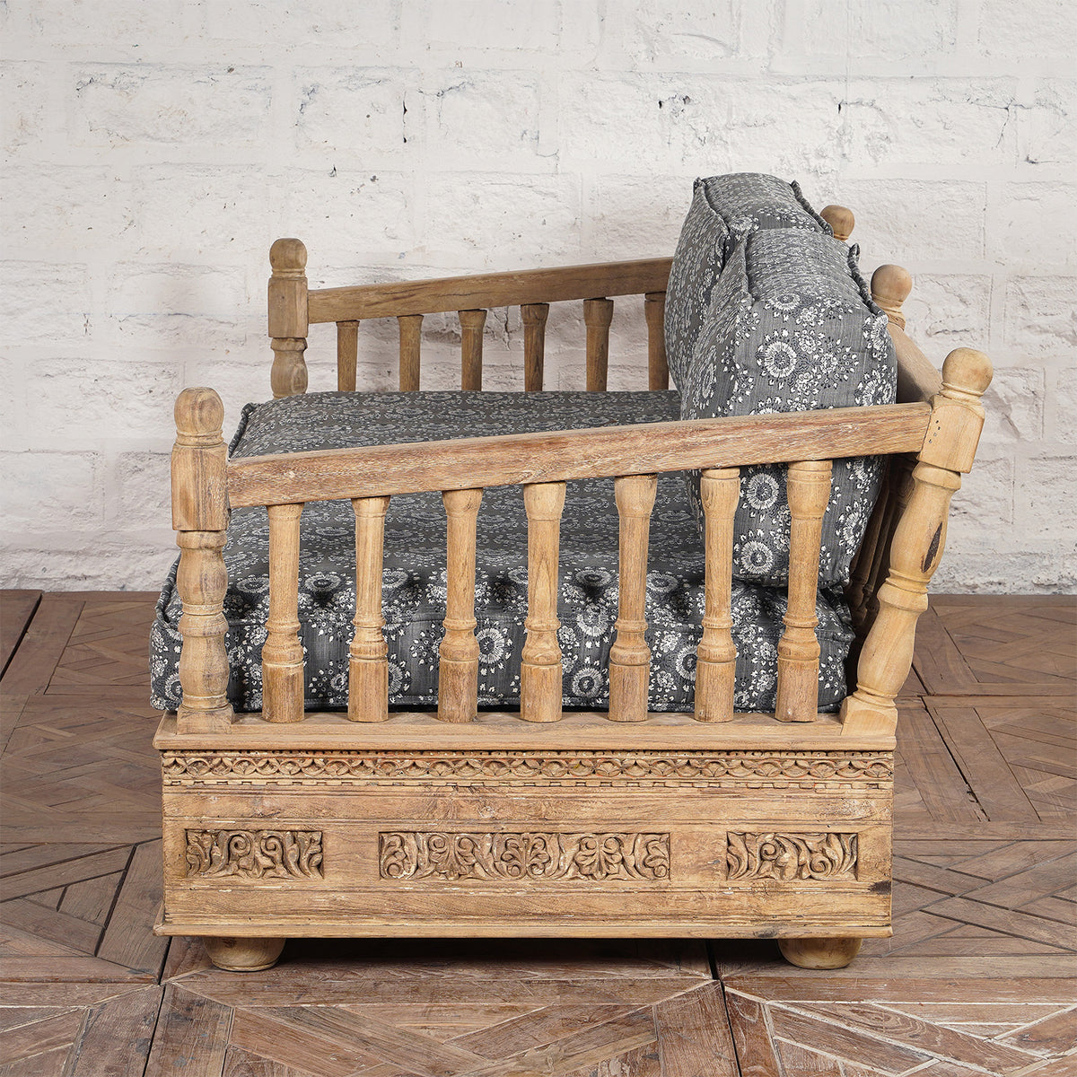 Side view of the Yashti Wooden Daybed by John Robshaw, featuring carved floral details, spindle armrests, and gray block printed fabric cushions, placed on a wooden floor against a textured white wall.