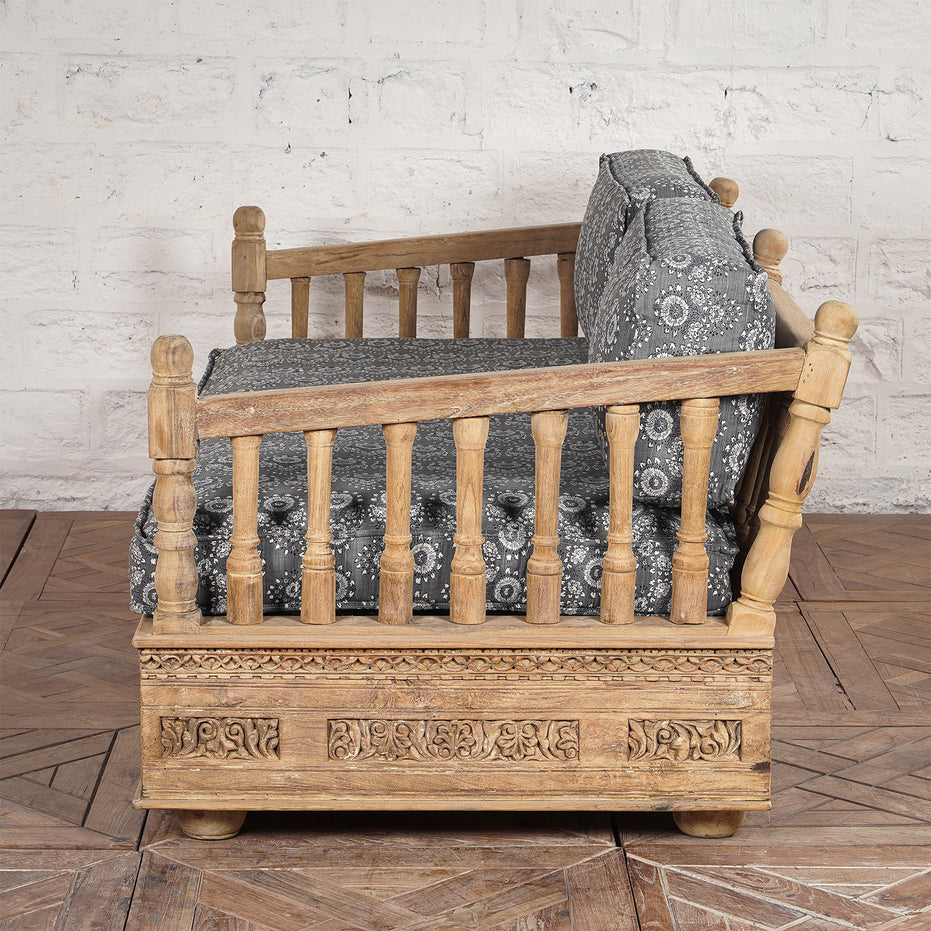 Side view of the Yashti Wooden Daybed by John Robshaw, featuring carved floral details, spindle armrests, and gray block printed fabric cushions, placed on a wooden floor against a textured white wall.