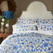 A cozy bedroom featuring a bed with a blue and green floral quilt and pillows, complemented by a patterned upholstered headboard in John Robshaw’s Mali Light Indigo Fabric, inspired by traditional Rajasthan flower designs created with handcarved blocks; accompanied by a side table with white flowers and a decorative lamp.