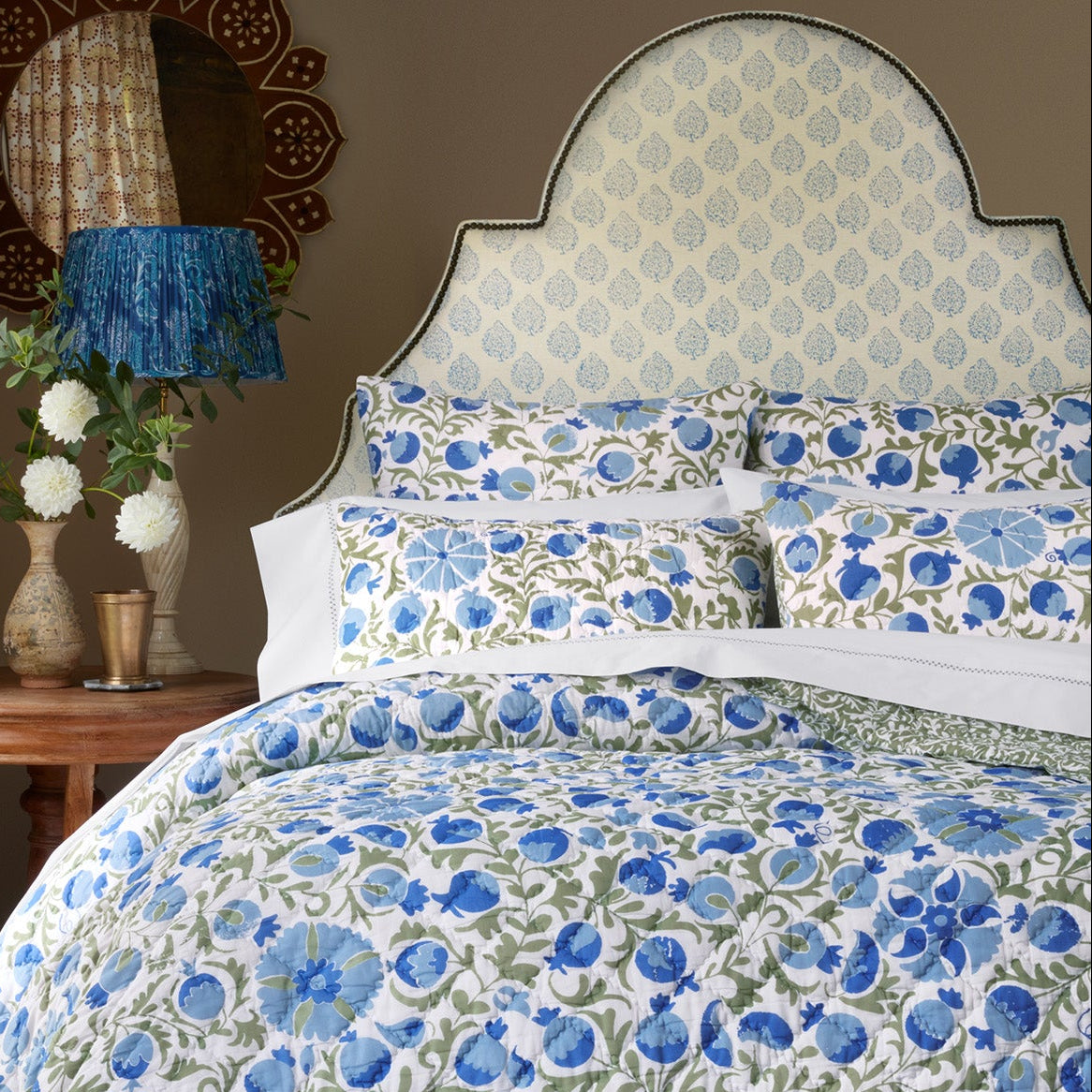 A cozy bedroom featuring a bed with a blue and green floral quilt and pillows, complemented by a patterned upholstered headboard in John Robshaw’s Mali Light Indigo Fabric, inspired by traditional Rajasthan flower designs created with handcarved blocks; accompanied by a side table with white flowers and a decorative lamp.