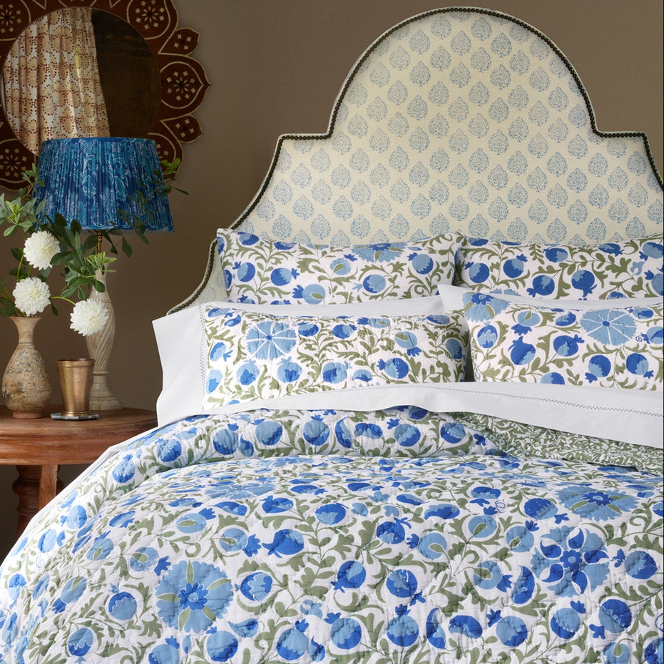 A cozy bedroom featuring a bed with a blue and green floral quilt and pillows, complemented by a patterned upholstered headboard in John Robshaw’s Mali Light Indigo Fabric, inspired by traditional Rajasthan flower designs created with handcarved blocks; accompanied by a side table with white flowers and a decorative lamp.