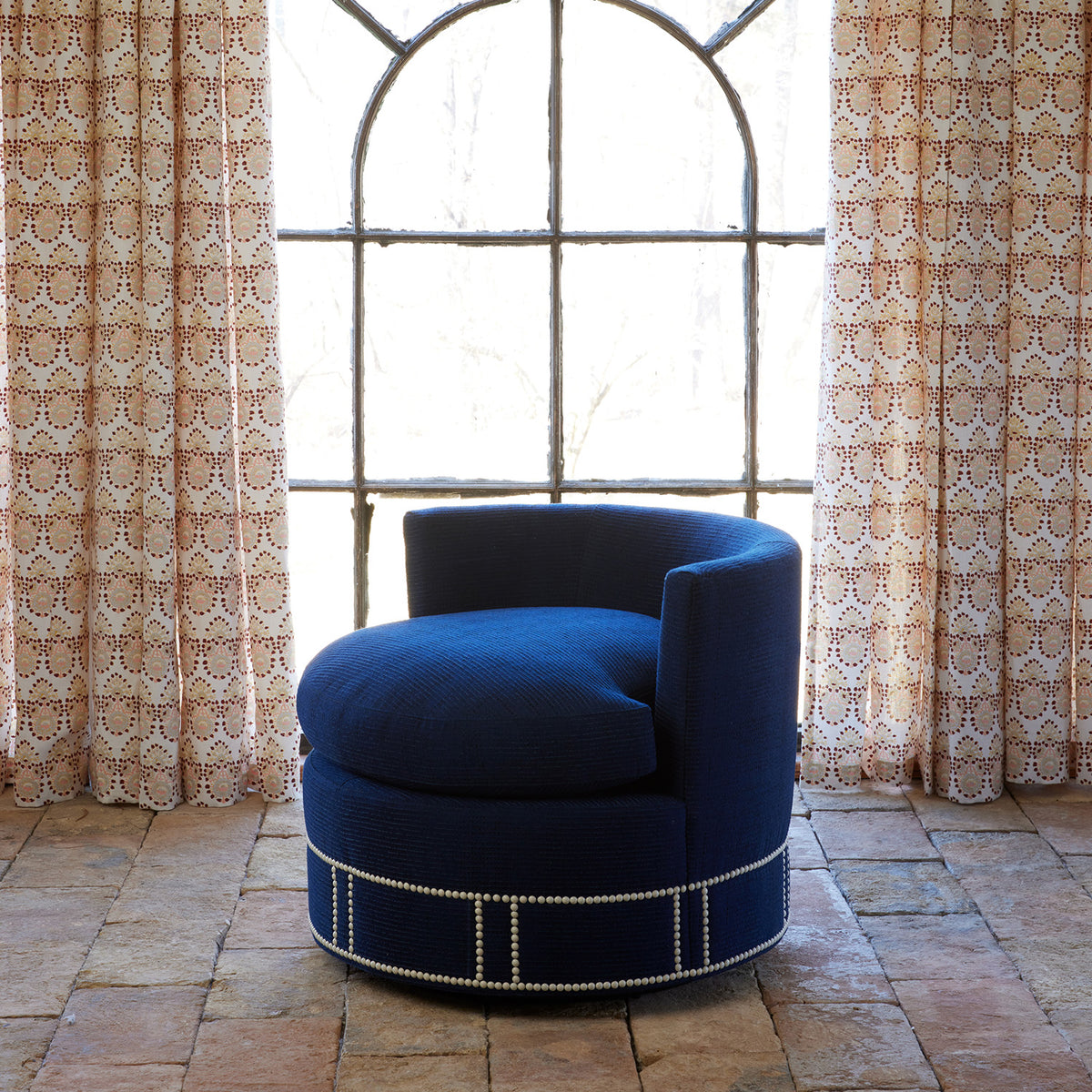 A Round Swivel Chair in Bijal Indigo by John Robshaw, featuring silver stud accents and upholstered in performance fabric, rests on a tiled floor before a large arched window draped with patterned curtains.