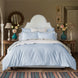 A neatly made bed featuring the John Robshaw Cinde Light Indigo Organic Duvet, flanked by wooden side tables holding vases of flowers and lamps, in a decorated bedroom.