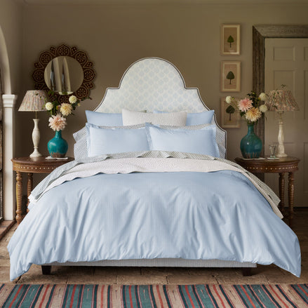A neatly made bed featuring the John Robshaw Cinde Light Indigo Organic Duvet, flanked by wooden side tables holding vases of flowers and lamps, in a decorated bedroom.