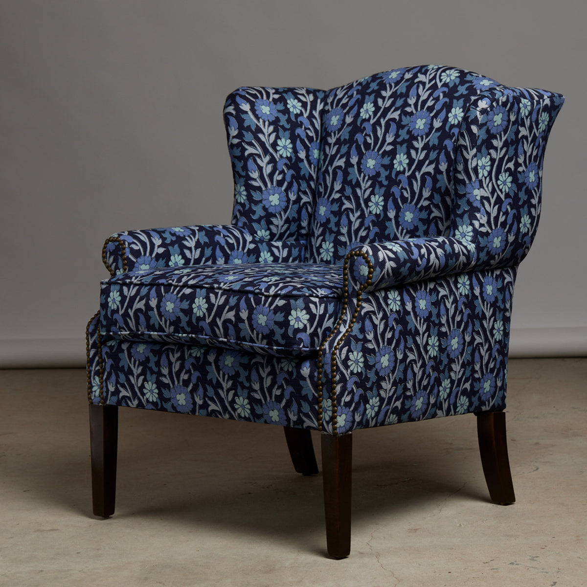 John Robshaw Ashi Azure Woodruff Chair with wooden legs, upholstered in Ashi Azure fabric showcasing a floral block print of light blue flowers and green leaves on a dark blue background.