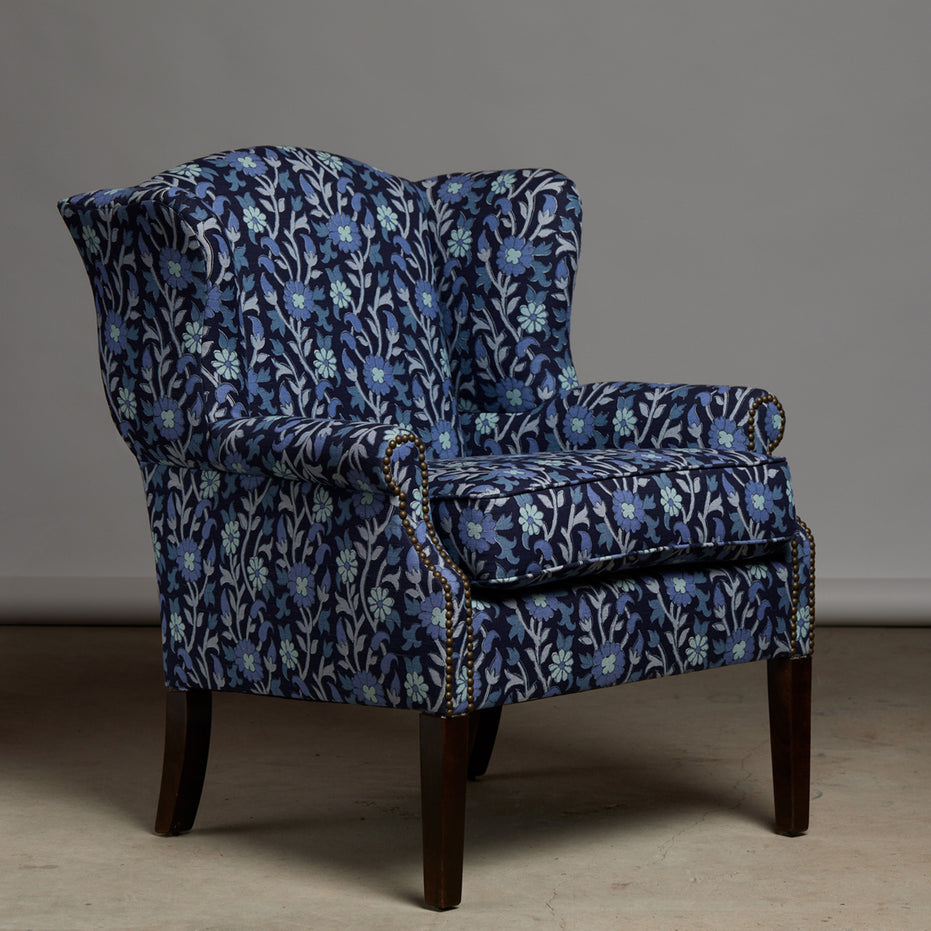 The John Robshaw Ashi Azure Woodruff Chair features elegant floral-patterned Ashi Azure fabric upholstery, dark wooden legs, and brass nailhead trim. Its classic wingback silhouette is enhanced by a sophisticated block print design and set against a plain gray background.