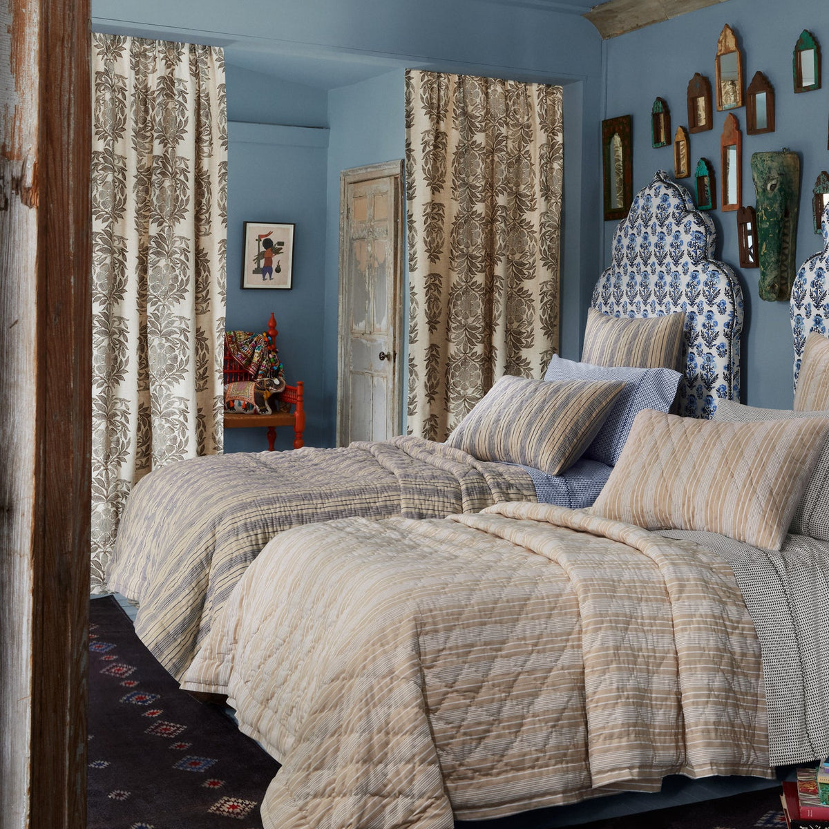 Bedroom with blue walls, striped bedding, dramatic curtains featuring a hand-drawn vine pattern, an arched headboard upholstered in John Robshaw’s Yashi Chestnut Fabric, and a gallery of small decorative mirrors on the wall.