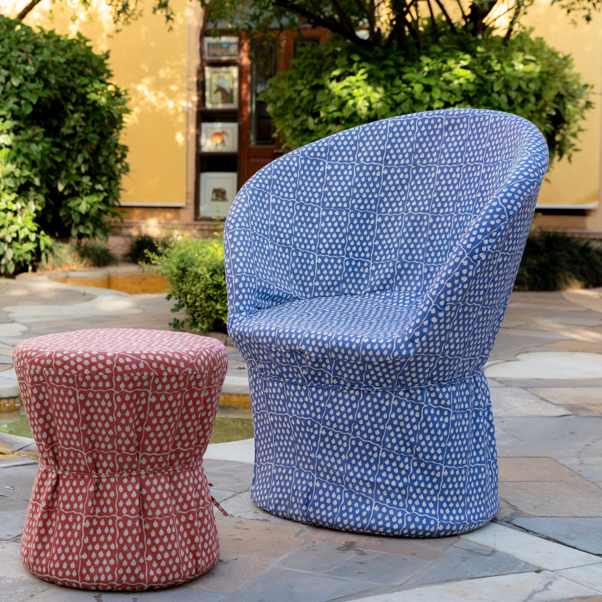 A chair upholstered in John Robshaw’s Balan Azure Fabric sits beside a red Sanganer clay print ottoman on a stone patio, surrounded by trees and bushes in the garden.