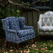 Two patterned armchairs, including the John Robshaw Ashi Azure Woodruff Chair featuring a delicate block-printed fabric, and another light chair with a paisley design, sit on the grass near a house with green siding and fallen leaves scattered on the ground.