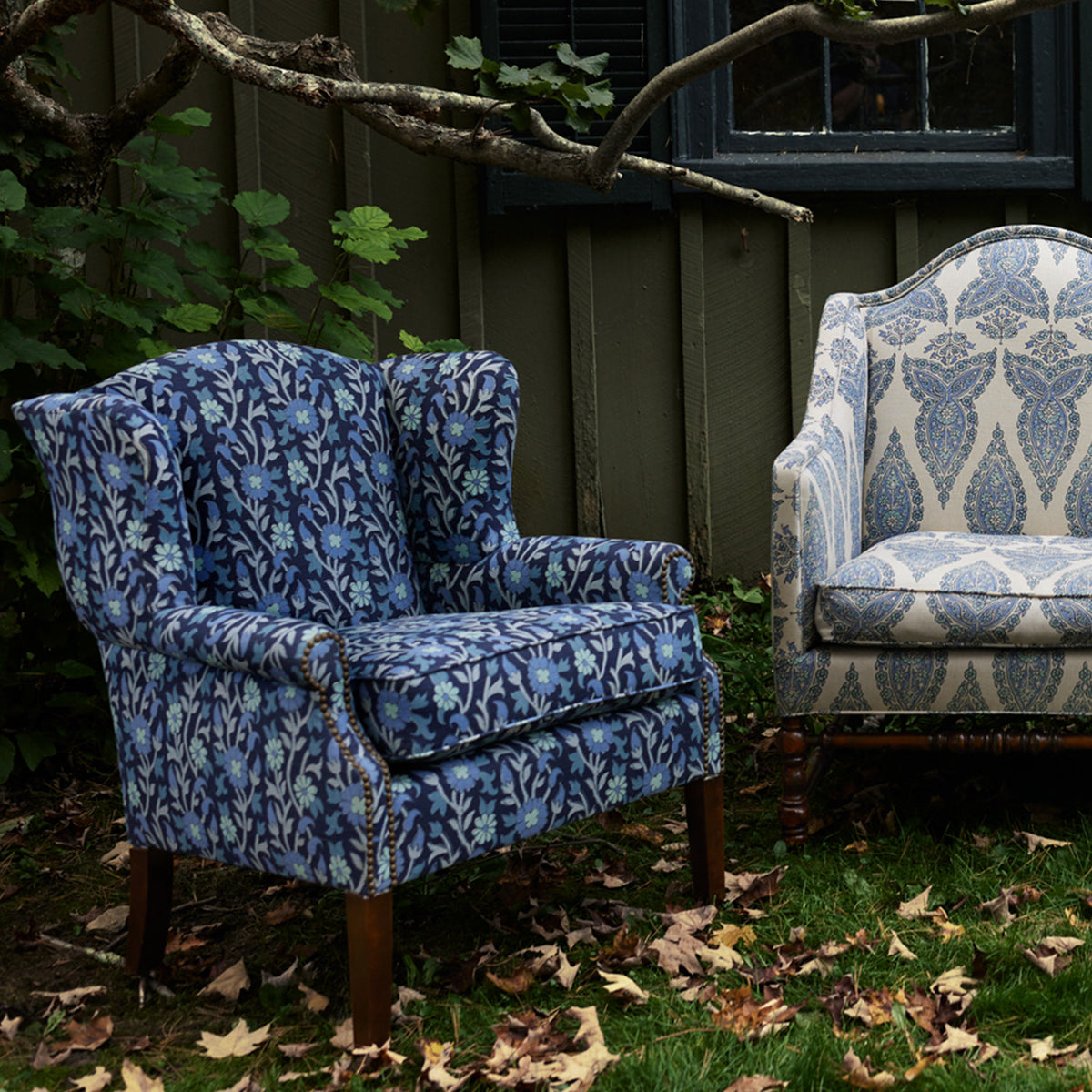 Two patterned armchairs, including the John Robshaw Ashi Azure Woodruff Chair featuring a delicate block-printed fabric, and another light chair with a paisley design, sit on the grass near a house with green siding and fallen leaves scattered on the ground.