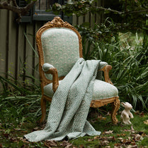 An ornate vintage-style wooden armchair with pale green patterned upholstery sits outdoors on grass beside a house wall, with a matching pale green throw blanket draped over the chair and a small white rabbit figurine nearby.