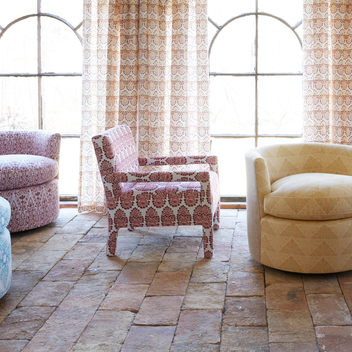 A room featuring John Robshaw’s Square Chair in Lanka Berry and Aleppo Berry performance fabric, paired with patterned armchairs on a brick floor, set before large arched windows draped with lace curtains.