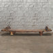 A low, rustic Wooden Naga Coffee Table 8 by John Robshaw—with short legs and a weathered surface evocative of antique tribal craftsmanship—is placed on a concrete floor against a light gray brick wall.