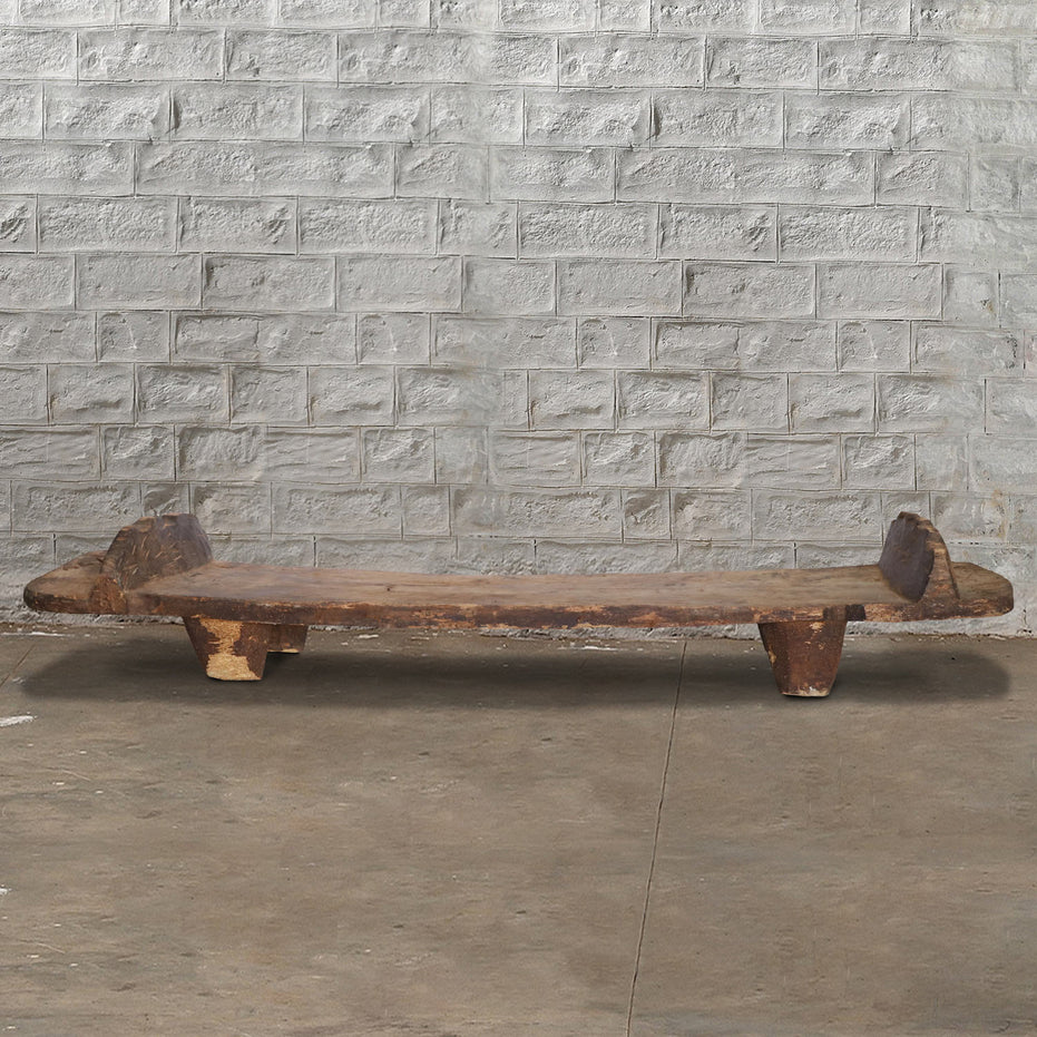 A low, rustic Wooden Naga Coffee Table 8 by John Robshaw—with short legs and a weathered surface evocative of antique tribal craftsmanship—is placed on a concrete floor against a light gray brick wall.