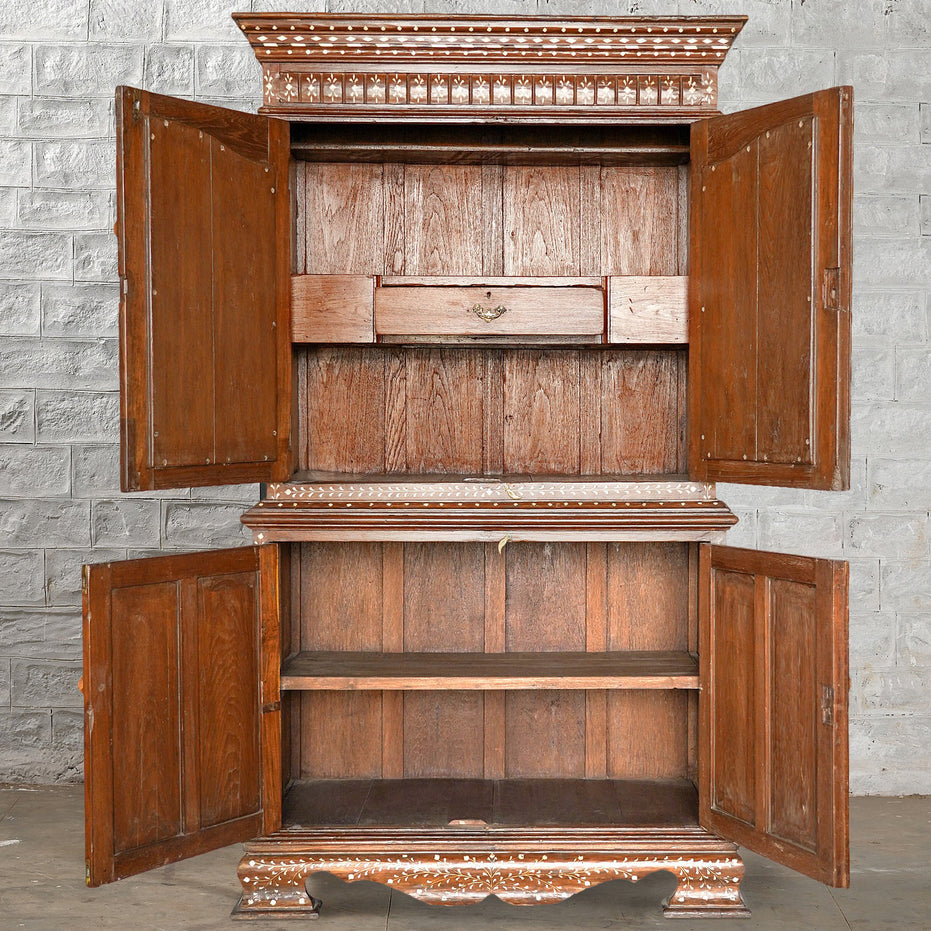 An Anglo Indian Teak Inlaid Cabinet by John Robshaw, featuring ornate trim, two shelves, and a small drawer, stands open against a gray brick wall—an exquisite piece evocative of classic bone inlay furniture.