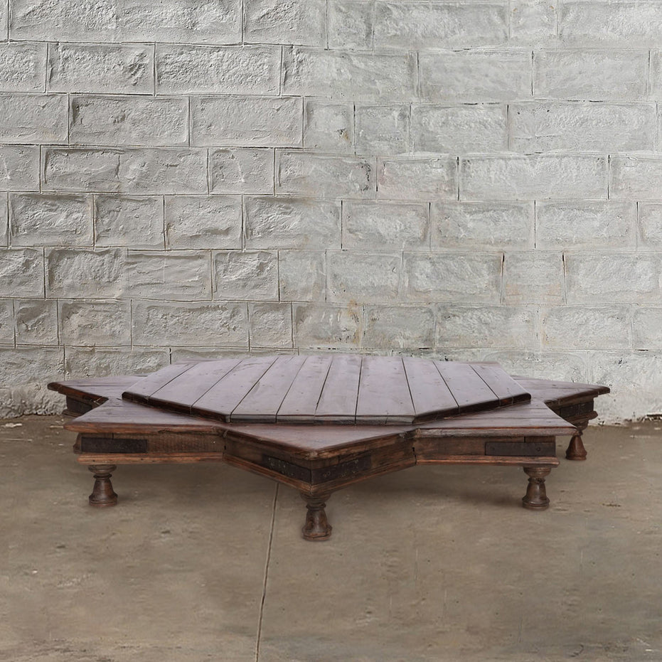 An 8 Point Wooden Bajot by John Robshaw, featuring a low, octagonal design inspired by vintage Rajasthani furniture and crafted from teak with short legs, is positioned against a white brick wall on a concrete floor.