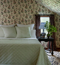 A styled bedroom scene featuring a bed with a light sage green quilt and matching pillows, set against a wall covered in vintage-style floral wallpaper. A dark wood nightstand with a blue-and-white ceramic lamp and a green potted plant stands beside the window.