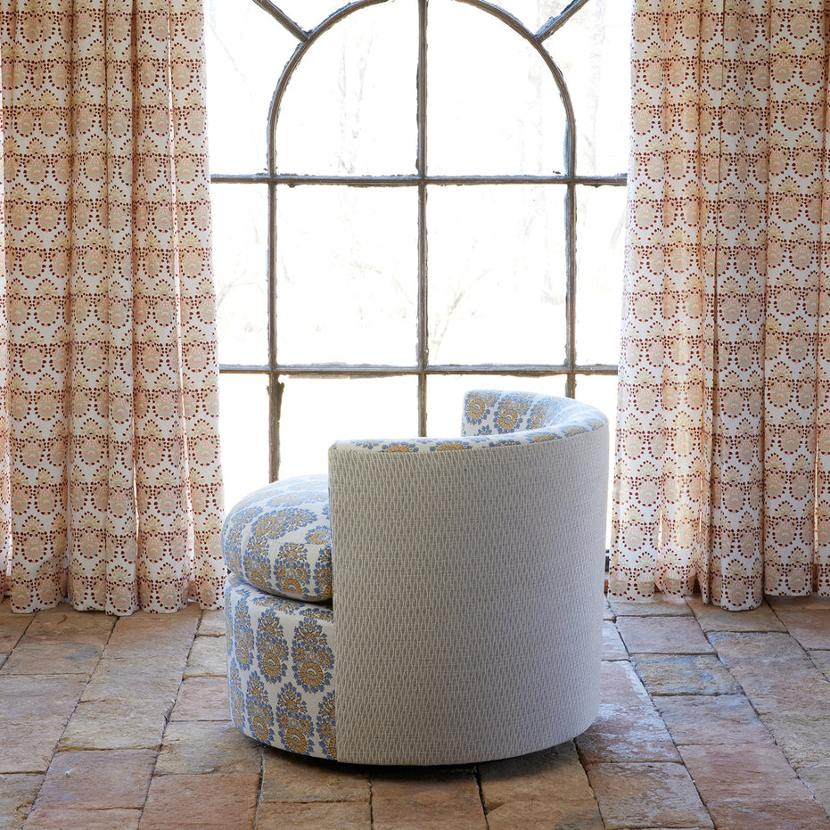 A Round Swivel Chair in Lanka Marigold and Natesh Gray by John Robshaw sits on a tiled floor in front of a large arched window with patterned curtains.
