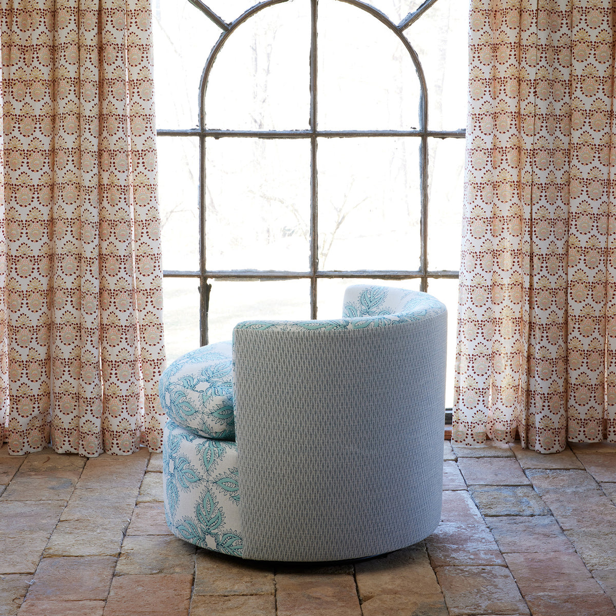A Round Swivel Chair in Diba Seaglass and Natesh Mist by John Robshaw is positioned on a stone floor in front of a tall arched window framed by patterned curtains.