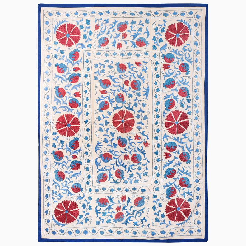 Jaipur Twilight Suzani Blanket: an off-white rectangular textile with blue and red botanical motifs in a traditional Suzani pattern and a solid blue border.