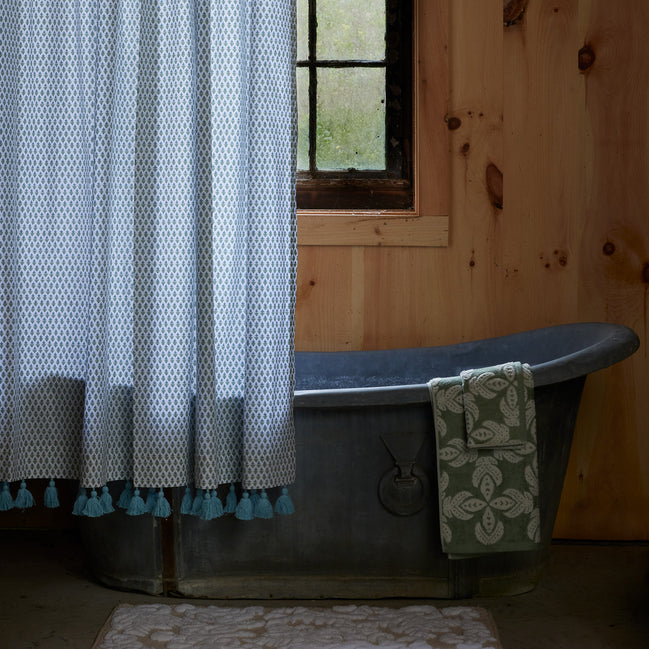 A metal bathtub with a patterned green towel, the John Robshaw Cala Sage Organic Shower Curtain in blue and white, and a wooden wall with a window in the background.