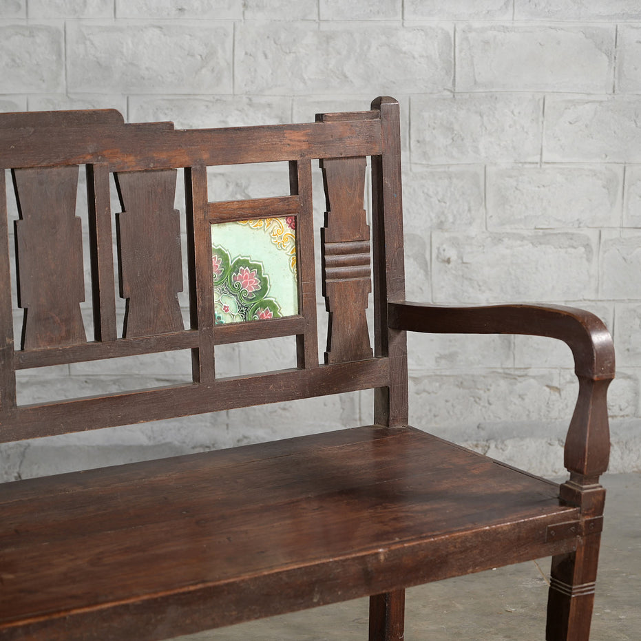 The Tipu Teak Bench by John Robshaw features a decorative backrest with a single tile showcasing a green floral pattern, and is set against a light gray brick wall.