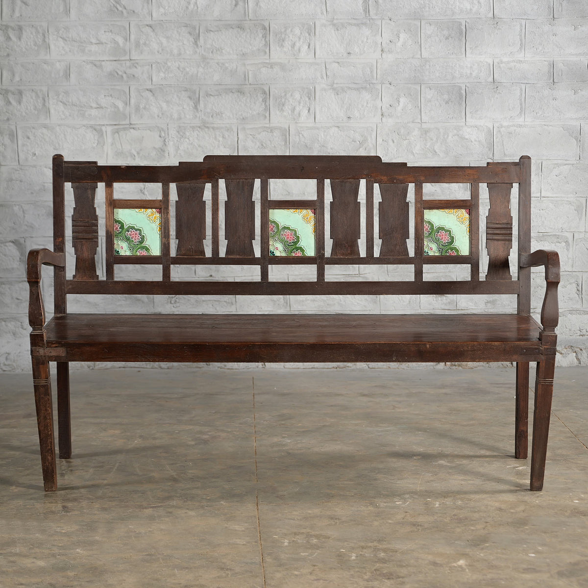 The Tipu Teak Bench by John Robshaw features a carved backrest and armrests, along with three decorative floral tile insets on the back. It is shown positioned against a light gray brick wall on a concrete floor.