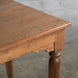 Detailed view of the corner of the Hanita Teak Table by John Robshaw, highlighting the finely turned legs and natural teak wood texture with visible joinery, set against a gray concrete wall background.
