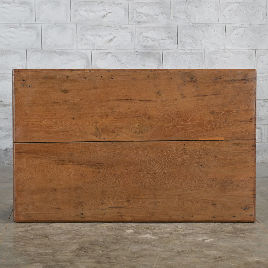 The Hanita Teak Table by John Robshaw, with its rectangular wooden design and smooth surface, is set against a textured white brick wall on a stone floor—an elegant piece of vintage furniture that brings timeless charm to any space.