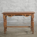 A handcrafted Daksh Teak Table by John Robshaw, featuring carved legs and decorative trim, is set against a light gray brick wall on a concrete floor.
