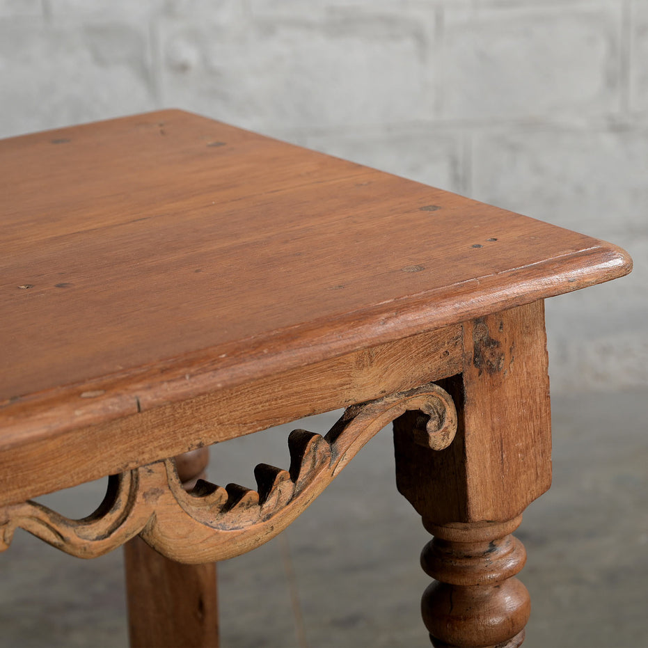 Close-up of the Daksh Teak Table by John Robshaw, showcasing its handcrafted corner with carved apron detailing and turned legs, set against a light-colored brick wall background—a refined example of vintage-inspired furniture.
