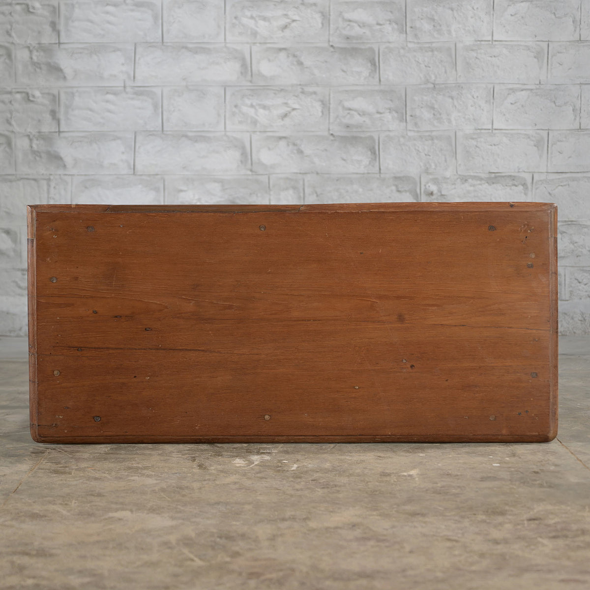 A Daksh Teak Table by John Robshaw sits on a concrete floor, set against a white brick wall and evoking the vintage charm and timeless appeal of handcrafted furniture.