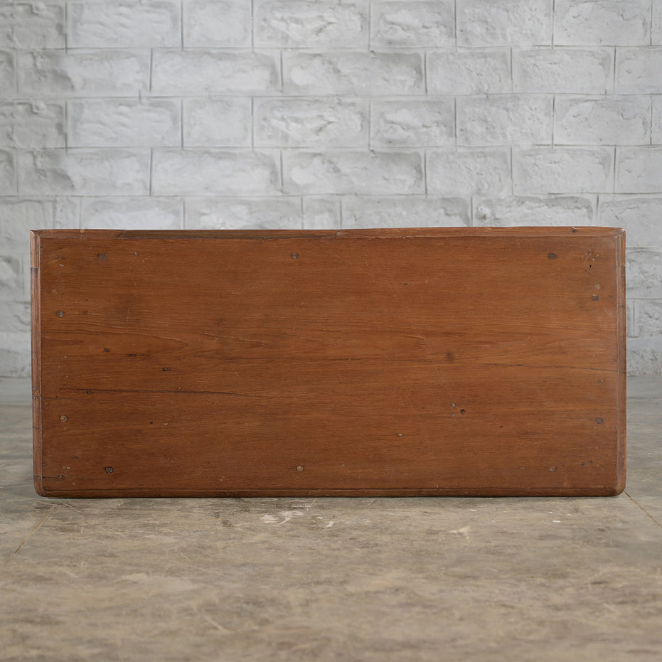 A Daksh Teak Table by John Robshaw sits on a concrete floor, set against a white brick wall and evoking the vintage charm and timeless appeal of handcrafted furniture.