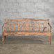 The Oorja Teak Bench by John Robshaw, featuring a handcrafted geometric backrest, stands on a concrete floor against a gray brick wall, its weathered wooden finish evoking the timeless appeal of vintage furniture.