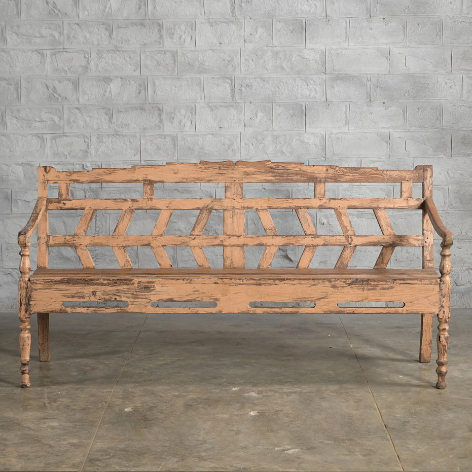The Oorja Teak Bench by John Robshaw, featuring a handcrafted geometric backrest, stands on a concrete floor against a gray brick wall, its weathered wooden finish evoking the timeless appeal of vintage furniture.
