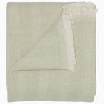 The Lavanya Sage Throw by John Robshaw is a folded, light cotton blanket featuring a subtle herringbone pattern and one fringed edge.