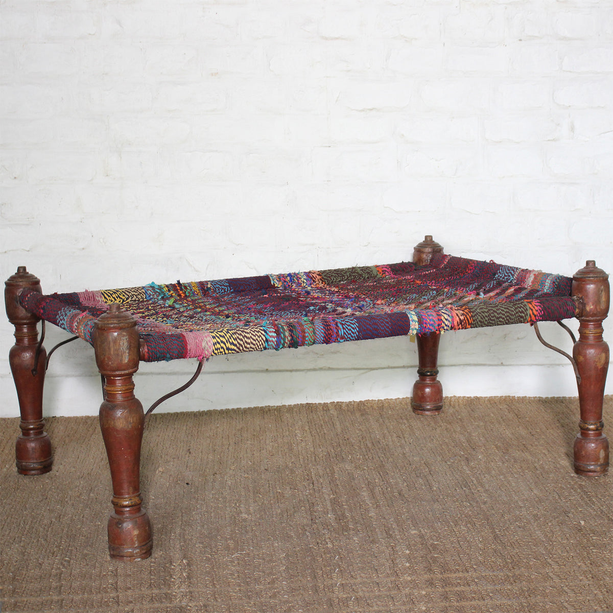 The Rangana Teak Charpoy by John Robshaw, featuring turned wooden legs and a vibrant surface woven from multicolored threads, stands on a woven mat against a white brick wall.