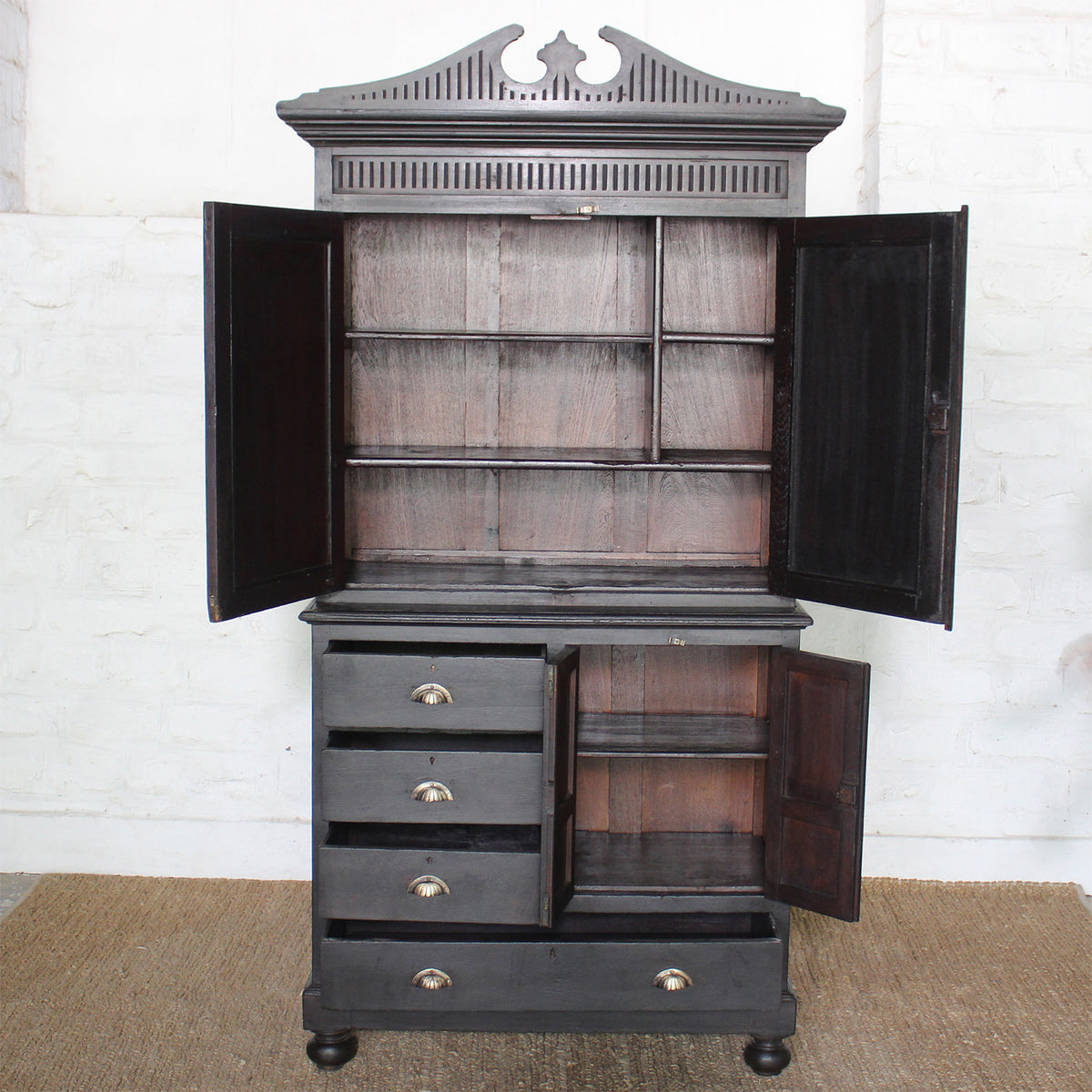 The Rajan Wooden Cabinet by John Robshaw features an ornate top and a black campaign-inspired design, with open upper and lower doors—one set boasting mirrored panels—that reveal versatile storage shelves, alongside four drawers with metal handles. It stands elegantly on a woven mat against a white brick wall.
