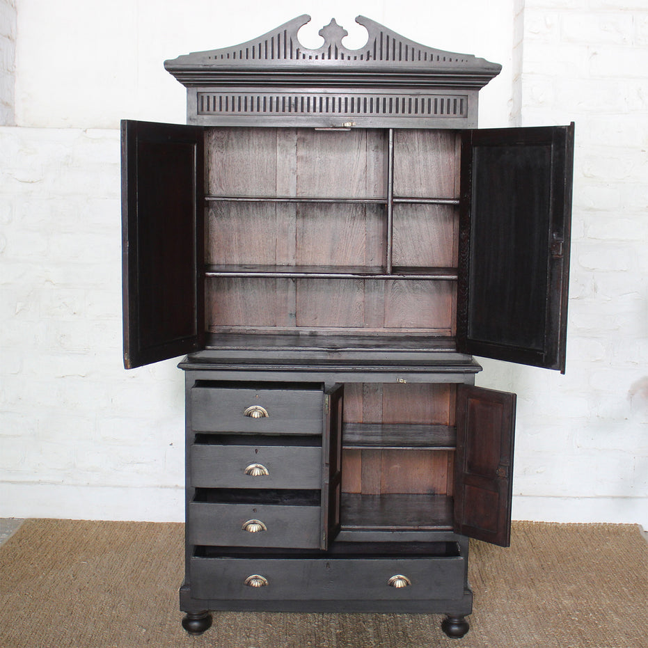 The Rajan Wooden Cabinet by John Robshaw features an ornate top and a black campaign-inspired design, with open upper and lower doors—one set boasting mirrored panels—that reveal versatile storage shelves, alongside four drawers with metal handles. It stands elegantly on a woven mat against a white brick wall.